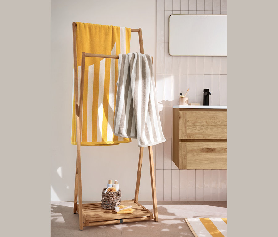 Ein gelb-weiß gestreiftes Jacquard-Badetuch und zwei beige-weiß gestreifte Handtücher hängen auf einem Klappbaren Kleiderständer. Darunter stehen 2 Aufbewahrungskörbe.