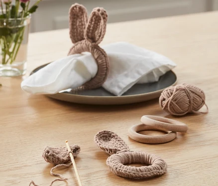 Auf einem Tisch liegt ein DIY-Häkelset »Serviettenringe«. Ein fertiger Ring mit Hasenohren liegt auf einer Serviette auf einem Teller. Daneben liegen ein angefangenes Häkelstück, ein Wollknäuel und Holzringe.