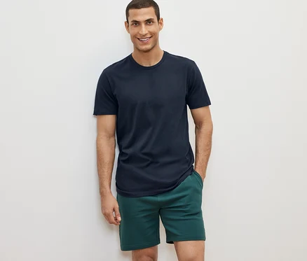 Mann posiert vor weißem Hintergrund in einem von 2 T-Shirts mit Rundhalsausschnitt, grüne Shorts.