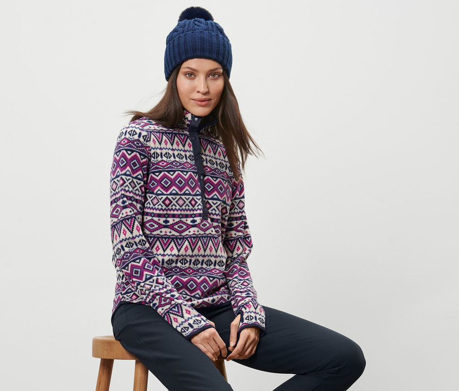 Frau sitzt auf einem Hocker und trägt einen Fleecepullover und eine blaue Mütze.