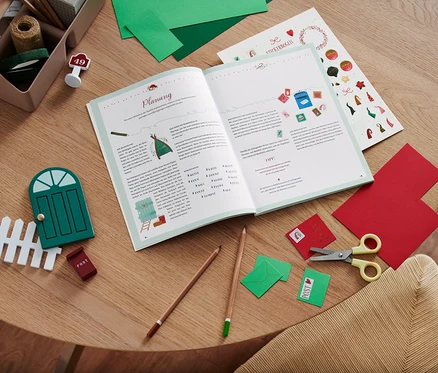 Auf einem Holztisch liegen ein offenes Buch, eine Schere, Buntstifte, bunte Papiere und Dekoartikel, inklusive das Buch »Hurra, ein Wichtel zieht ein!«.