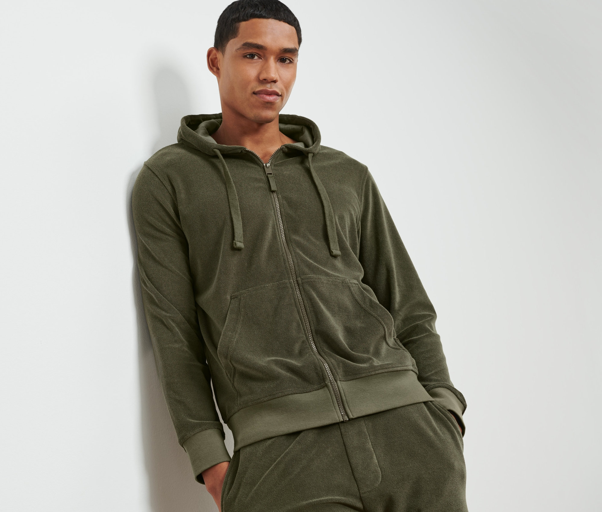 Mann lehnt an einer Wand und trägt eine Frottee-Kapuzenjacke.