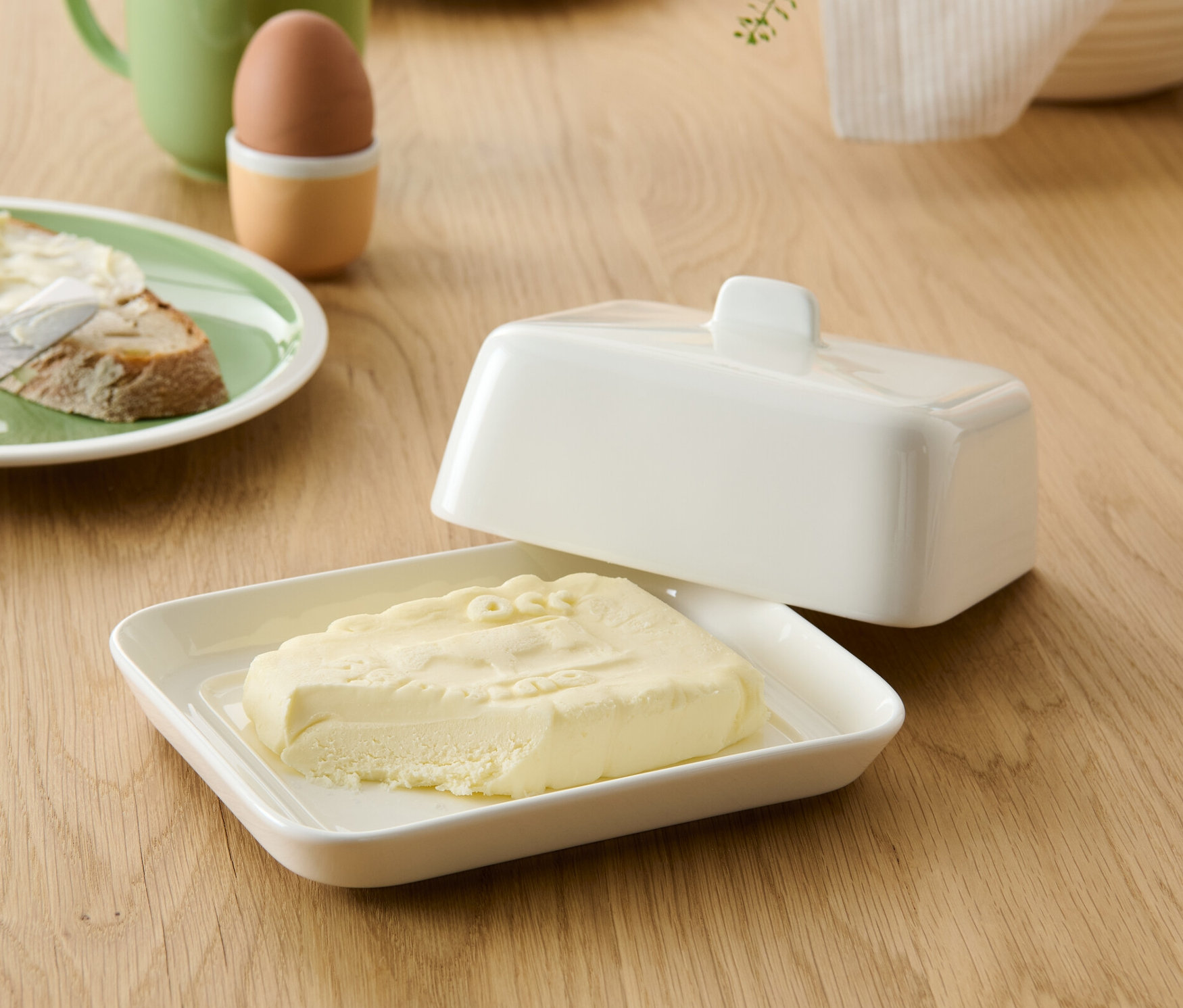Butter in offener Butterdose neben einem Butterbrot, im Hintergrund eine Tasse und ein Ei.