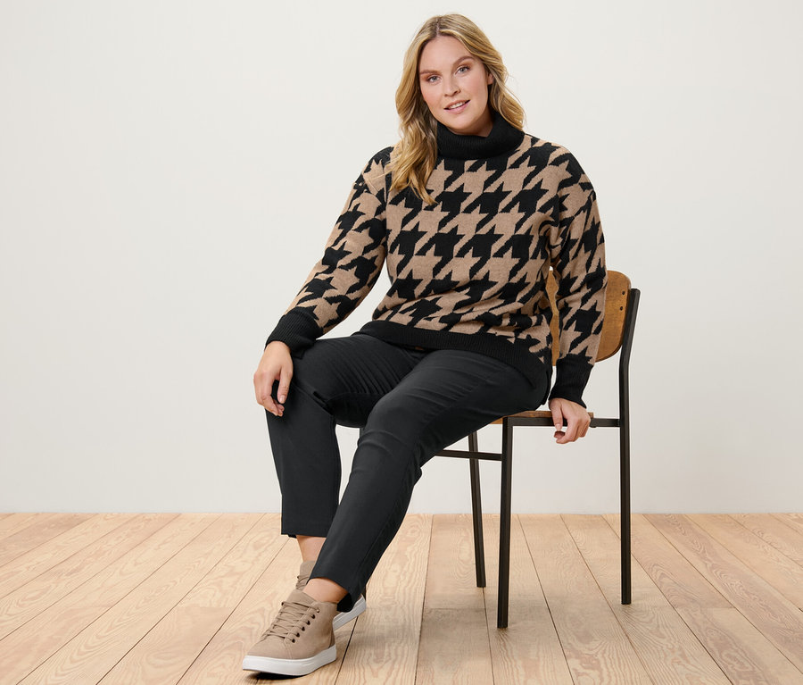 Frau sitzt auf einem Stuhl und trägt einen Strickpullover mit Hahnentritt-Jacquard und eine Stretchhose.