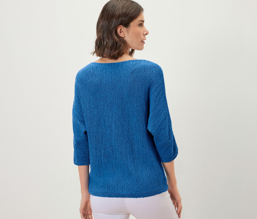 Frau posiert mit dem Rücken zur Kamera und trägt einen blauen sommerlichen Strickpullover aus Bändchengarn und weiße Hosen.