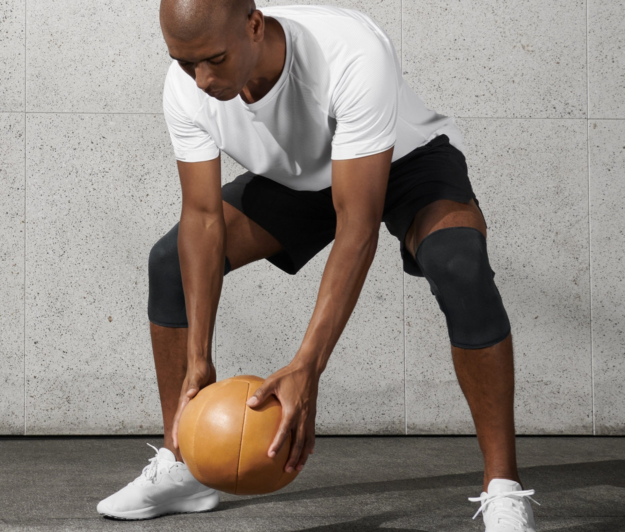 Mann in weißem T-Shirt und schwarzen Shorts hält Medizinball und trägt 2 Fitness-Knie-Manschetten.