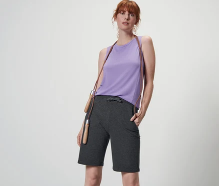 Frau posiert in anthrazitfarbenen Funktionsshorts und lila Top mit einem Springseil um den Hals.