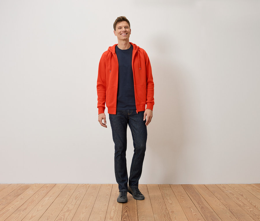 Ein lächelnder Mann steht mit orangefarbenem Hoodie, dunkelblauem T-Shirt und Jeans.
