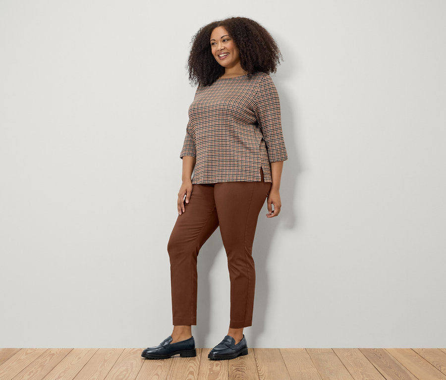 Frau posiert in braunem Sweatshirt mit 3/4-Arm, brauner Hose und schwarzen Schuhen.