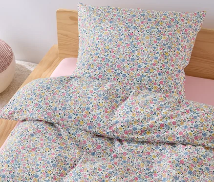 Auf einem Bett liegt Kinder Renforcé-Bettwäsche in Normalgröße mit Blumenmuster, daneben steht ein rosa Pouf.