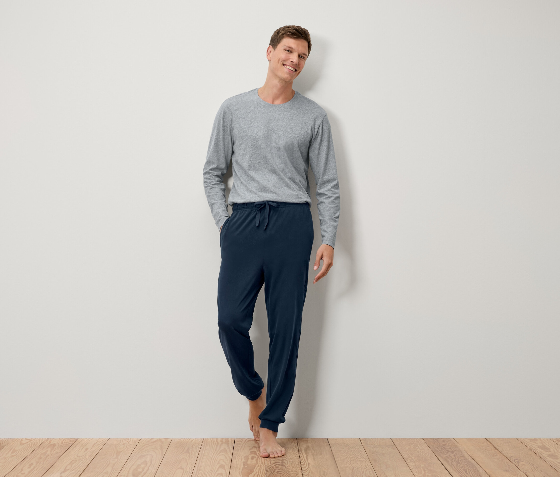 Ein barfüßiger Mann steht an einer Wand in einem grauen Langarmshirt und einer marineblauen Jogginghose.