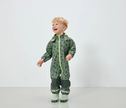 Ein Junge im grünen Overall mit Fleckenmuster und grünen Gummistiefeln springt in eine Pfütze.