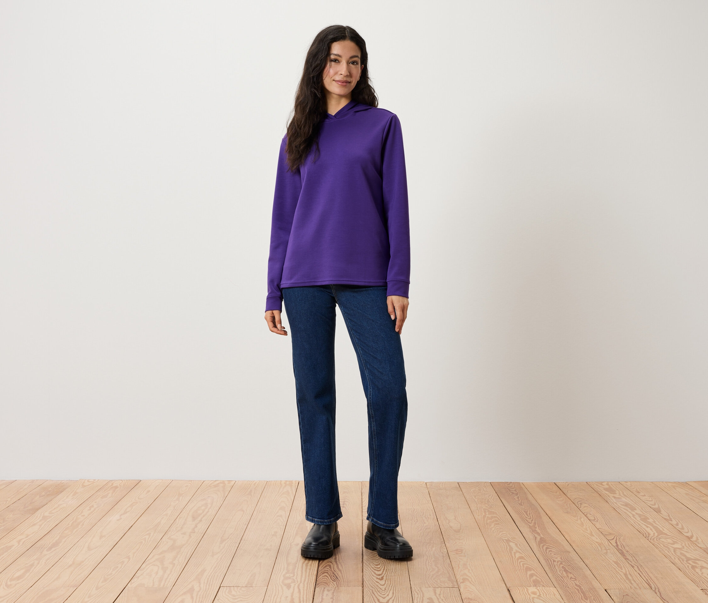 Eine Frau steht und trägt einen violetten Kapuzenpullover, Jeans und schwarze Stiefel.