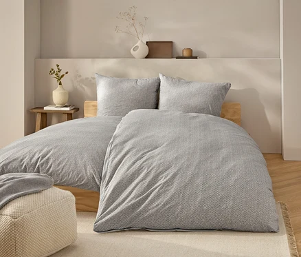 Bett mit grauer Bettwäsche und zwei Kissen. Auf dem Bett liegt eine graue Decke. Neben dem Bett steht ein beiger Pouf.