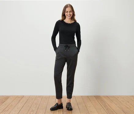 Eine Frau steht und lächelt, sie trägt ein schwarzes Langarmshirt, eine graue gemusterte Hose und schwarze Slipper.