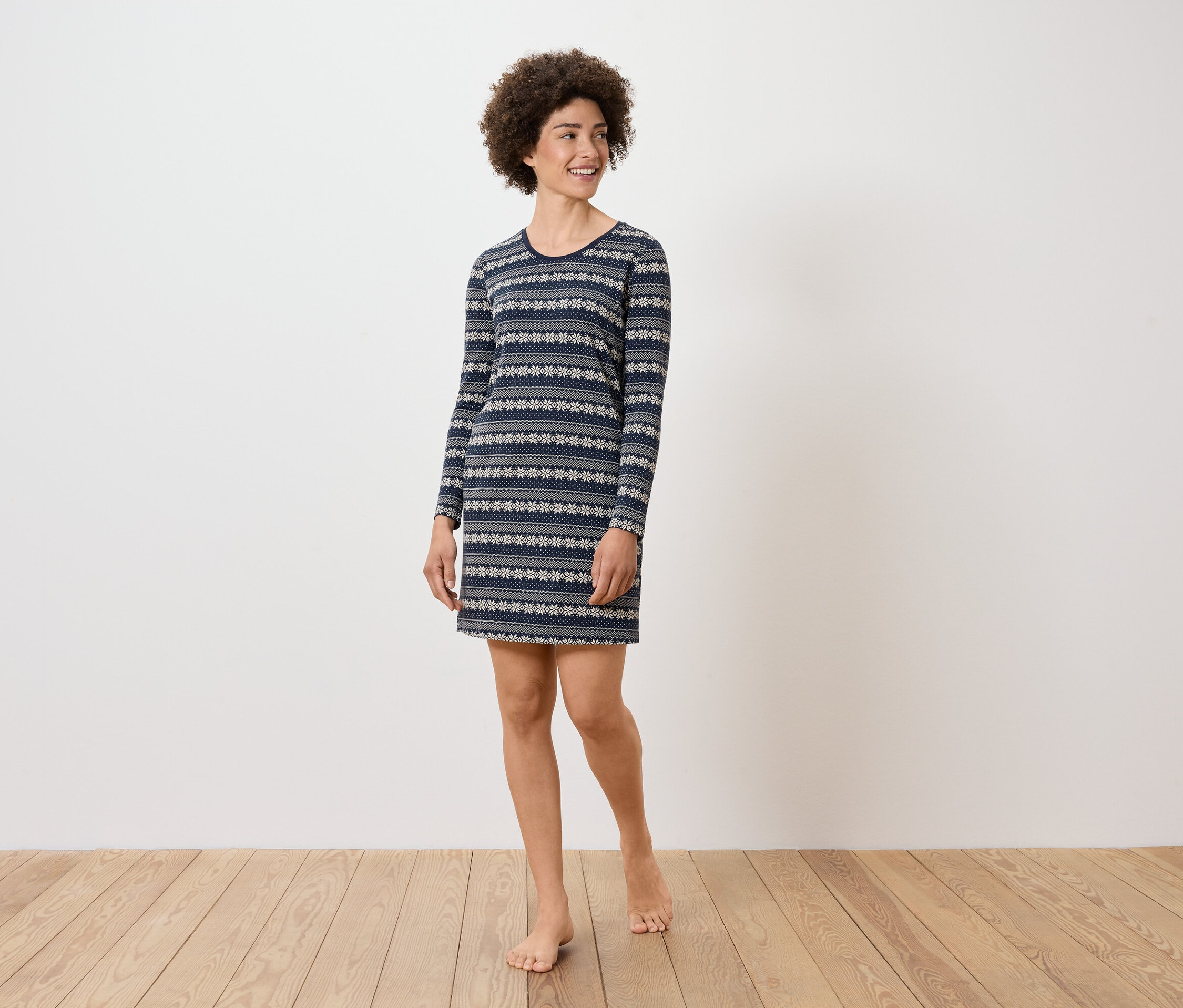 Frau mit lockigem Haar steht und trägt ein blaues, langärmeliges Kleid mit Muster.