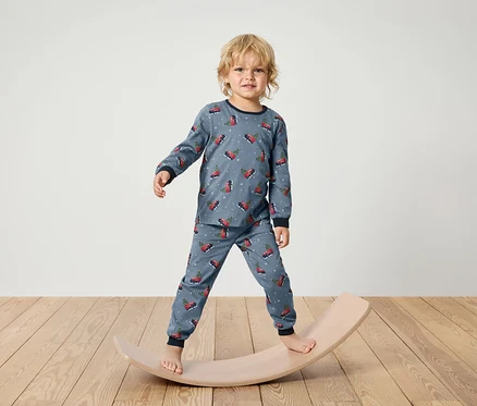 Ein Junge im blauen Schlafanzug mit Weihnachtsmotiv steht auf einem hölzernen Balance Board.