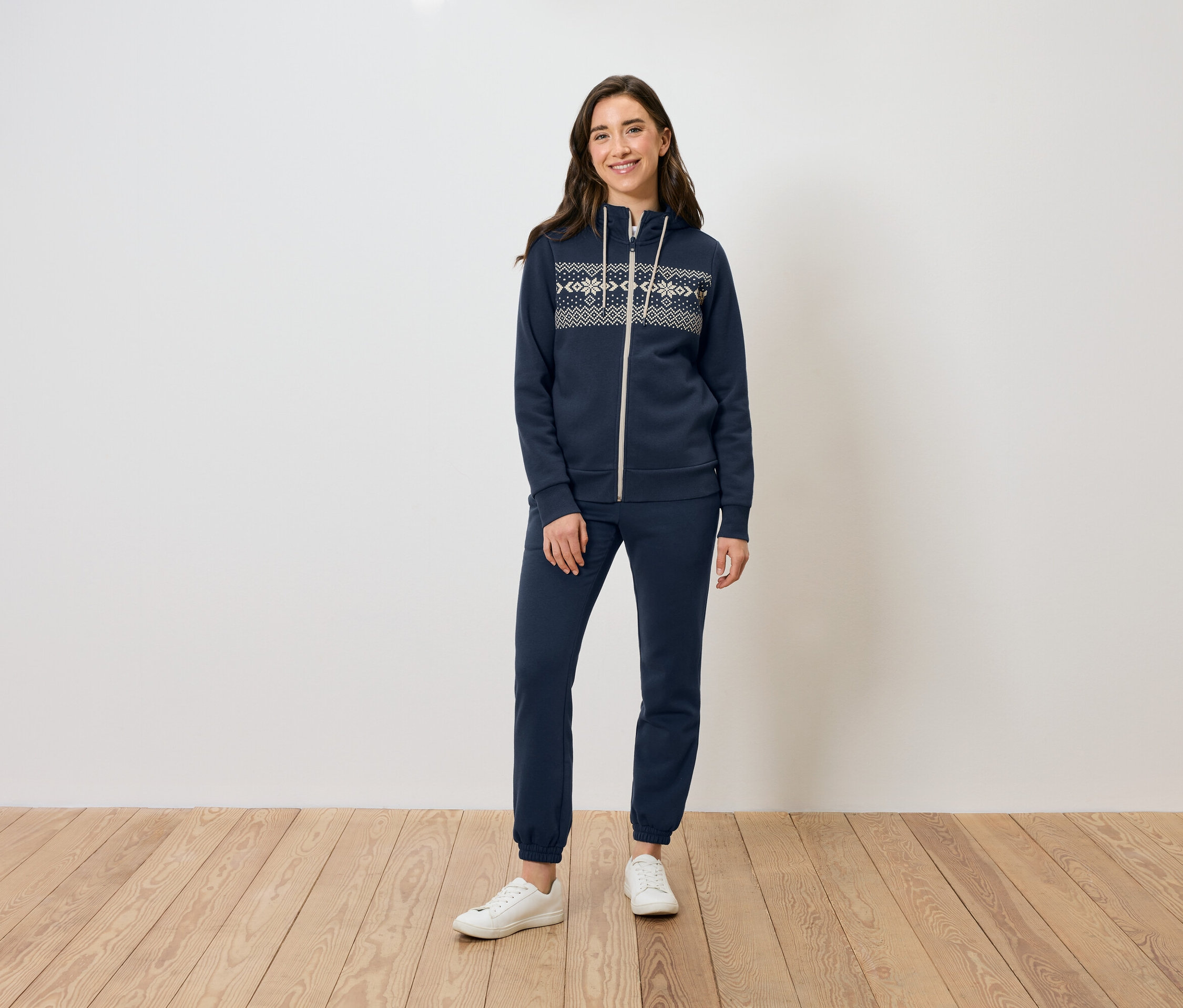 Eine Frau steht auf einem Holzboden und trägt einen dunkelblauen Reißverschluss-Hoodie und eine Trainingshose sowie weiße Turnschuhe.