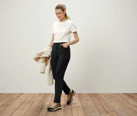 Frau geht auf einem Holzboden, trägt eine schwarze Hose, ein weißes T-Shirt und braune Schuhe. Sie hält einen cremefarbenen Pullover in der Hand.