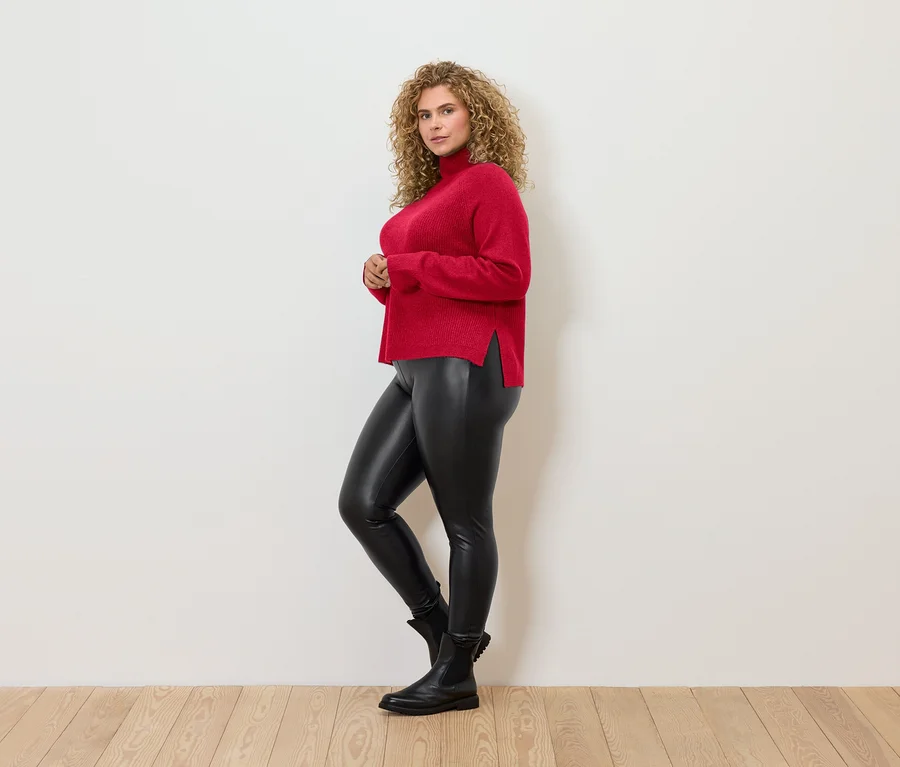Frau mit lockigem Haar posiert seitlich und trägt einen roten Rollkragenpullover, schwarze Leggings und schwarze Stiefel.
