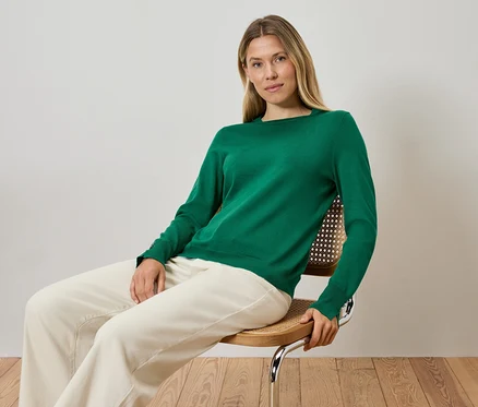 Blonde Frau sitzt auf einem Stuhl, trägt einen grünen Pullover und cremefarbene Hosen.