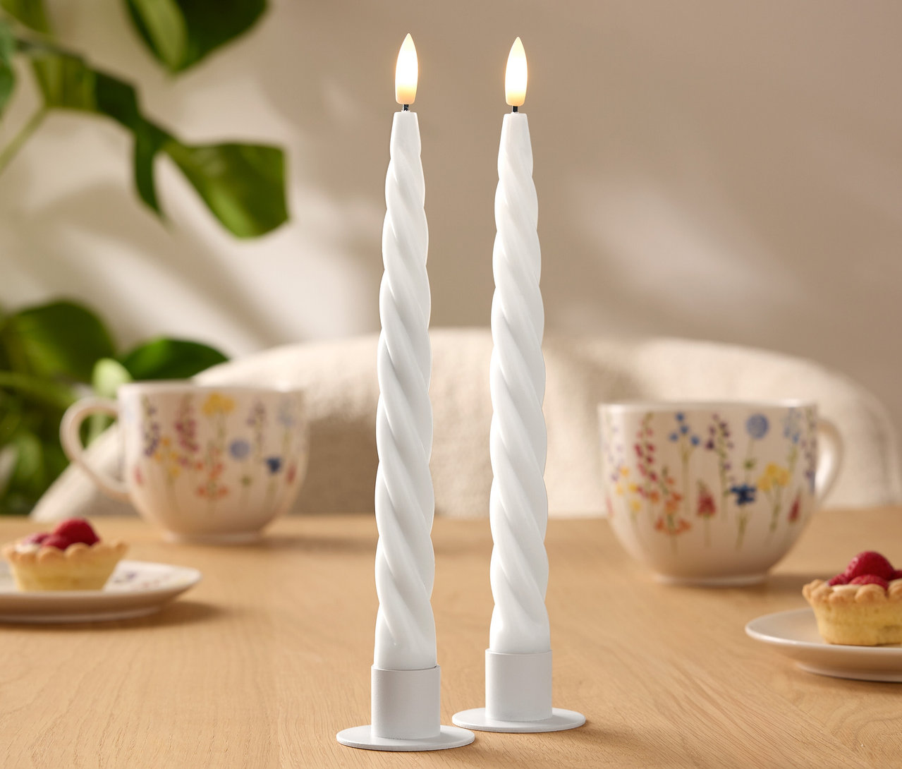 Zwei weiße, spiralförmige LED-Kerzen stehen auf einem Holztisch. Im Hintergrund sind zwei Tassen mit Blumenmuster und kleine Törtchen zu sehen.