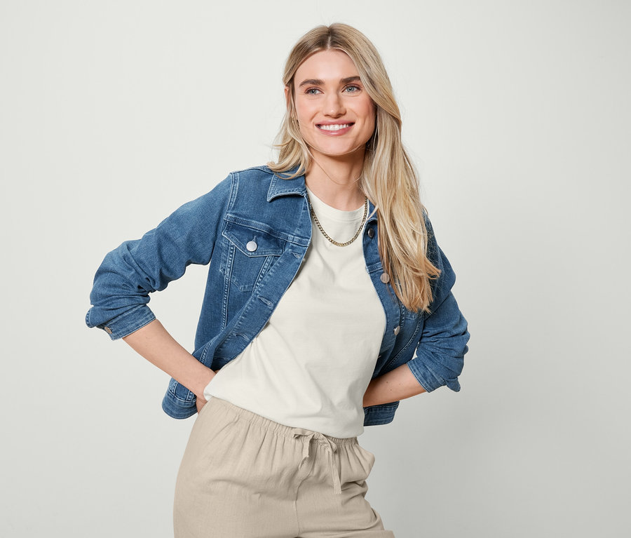 Eine lächelnde blonde Frau posiert in Jeansjacke, weißem Basic T-Shirt und sandfarbener Leinenhose.