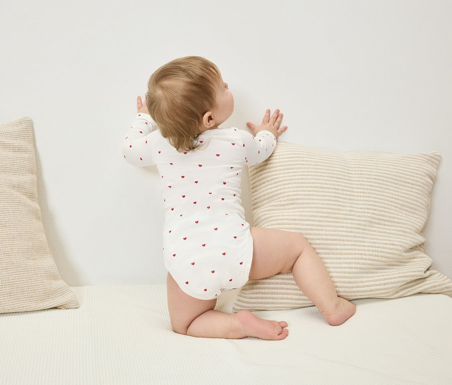 Kleinkind in einem langärmeligen Body mit roten Herzen, das an einer Wand neben einem gestreiften Kissen steht.