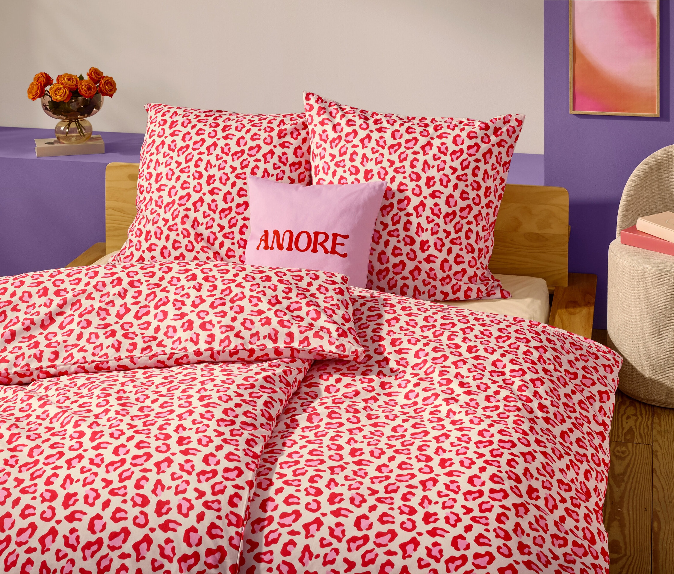 Nahaufnahme eines Bettes mit rot-weißem Leopardenmuster-Bettbezug und Kissen. Ein lavendelfarbenes Kissen mit dem Wort "AMORE" darauf befindet sich vor zwei Kopfkissen.