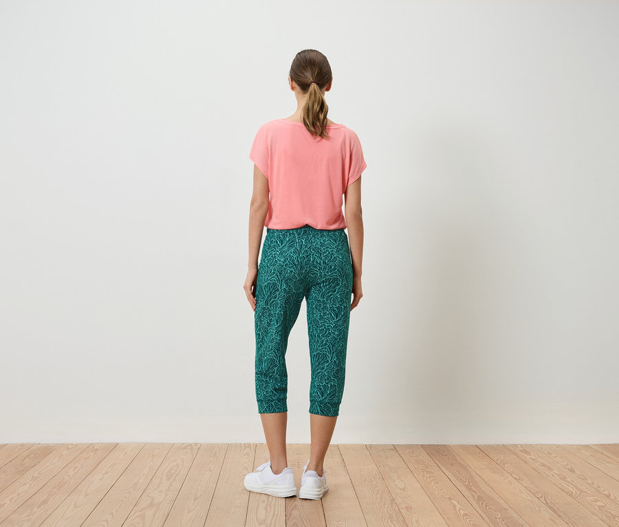 Frau von hinten in korallfarbenem T-Shirt und grüner Caprihose mit Blätterprint, dazu weiße Turnschuhe.