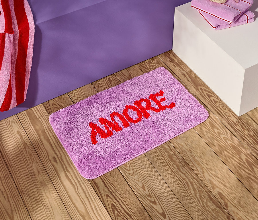 Ein lila Badezimmerteppich mit dem roten Wort "AMORE" liegt auf einem Holzboden neben einem lila Nachttisch.
