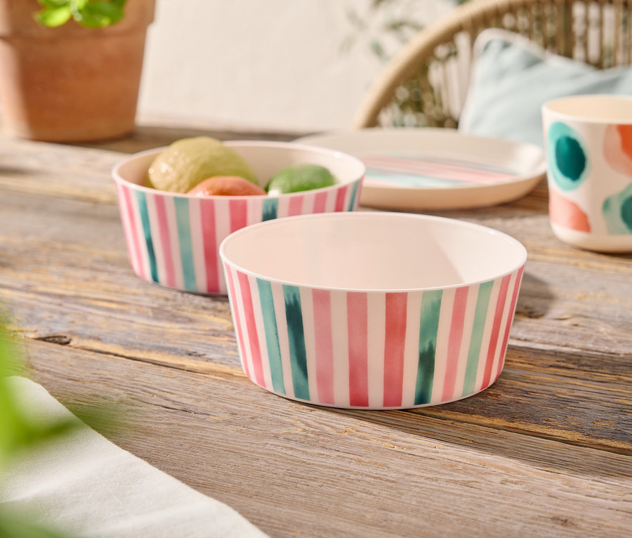 Auf einem Holztisch stehen zwei gestreifte Schüsseln, eine davon gefüllt mit Obst, daneben ein Teller, eine Tasse mit bunten Punkten und eine Topfpflanze.
