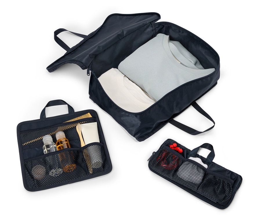 Drei geöffnete, marineblaue Reisetaschen mit Griffen und Reißverschlüssen liegen aufgereiht auf einem weißen Hintergrund. Die Taschen enthalten Kleidung, Kosmetika und Zubehör.