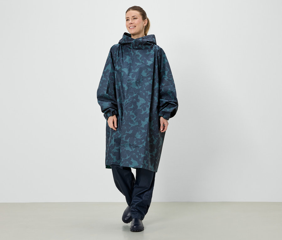 Eine Frau steht in einem langen, blauen Regenponcho mit Kapuze und einem Blattmuster.