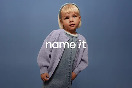 Kleines Kind mit blonden Haaren und blauem Haarband trägt eine Jeanslatzhose und eine lila Strickjacke vor einem blauen Hintergrund.