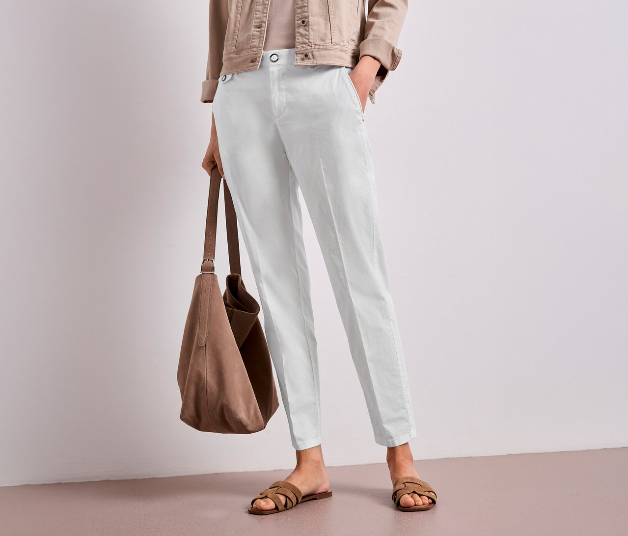Eine Person trägt eine hellbraune Jacke, ein helles Oberteil und eine weiße Street One Chino Hose, hält eine braune Wildledertasche und trägt braune Sandalen. Eine Hand steckt in der Hosentasche.