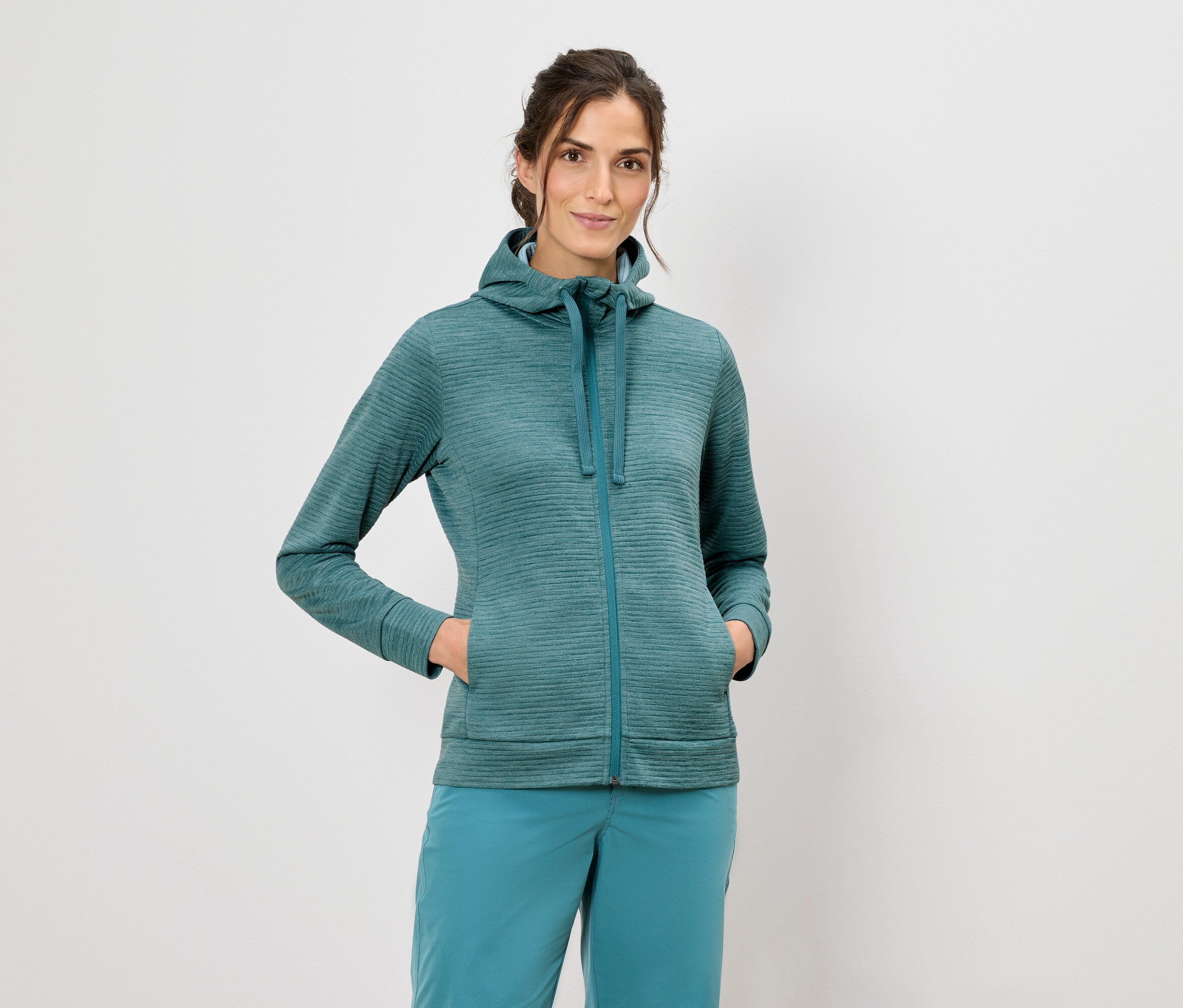 Frau mit Kapuzenjacke und blauer Hose steht vor einem weißen Hintergrund.