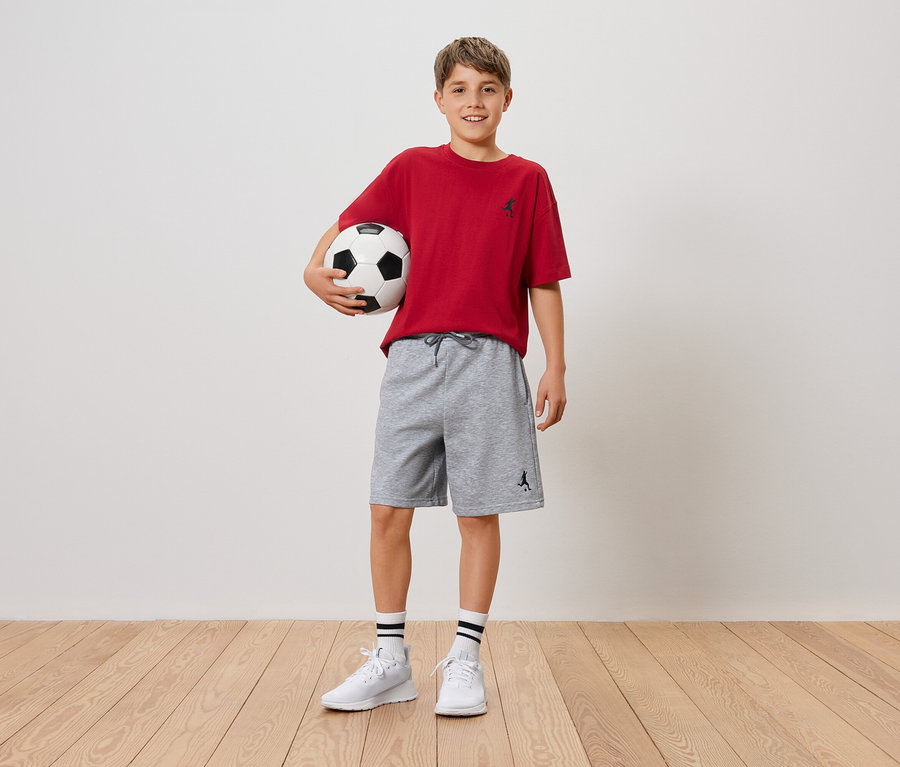 Ein lächelnder Junge hält einen Fußball in seinen Armen, er trägt ein rotes T-Shirt und graue Shorts.