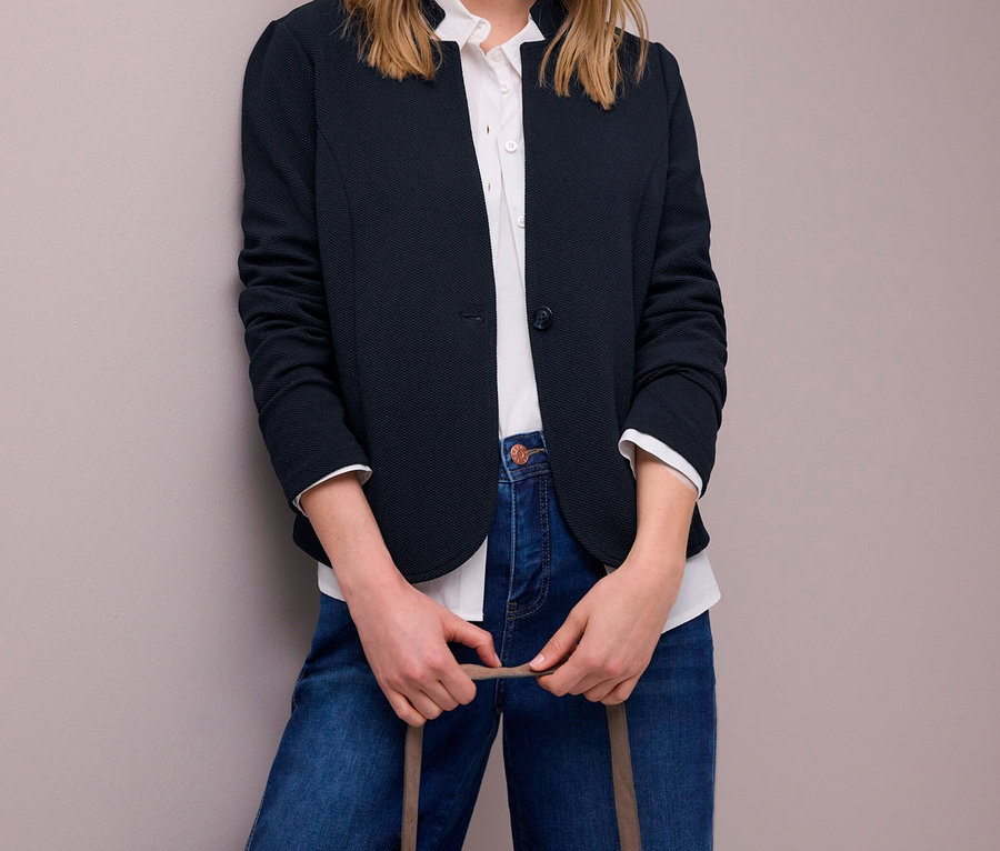 Blonde Frau trägt einen dunkelblauen Street One Jerseyblazer, ein weißes Hemd und blaue Jeans, eine hellbraune Handtasche haltend.