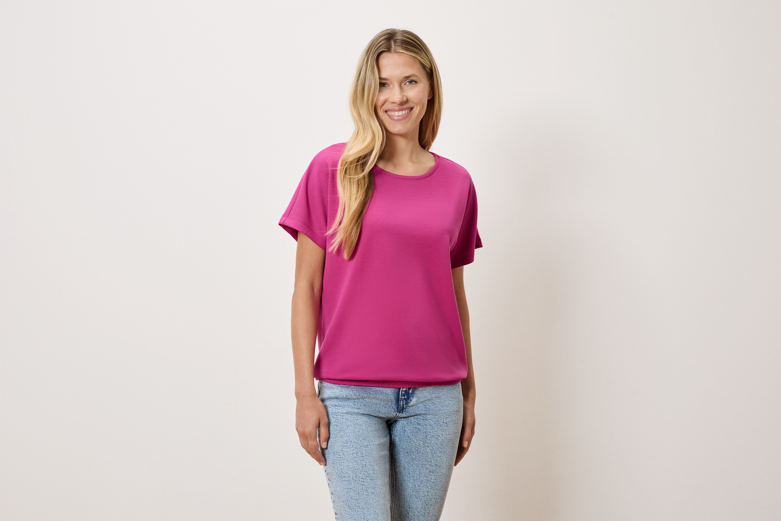 Eine Frau mit blonden Haaren trägt ein pinkes T-Shirt und blaue Jeans.