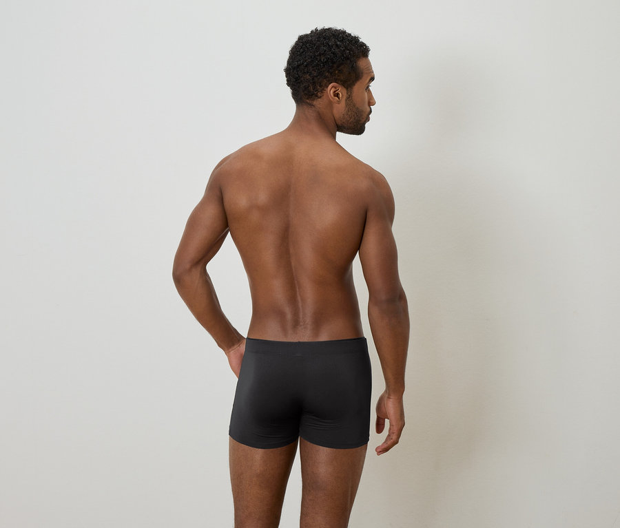 Afroamerikanischer Mann von hinten, mit nacktem Oberkörper und schwarzer Unterwäsche vor einem hellen Hintergrund.