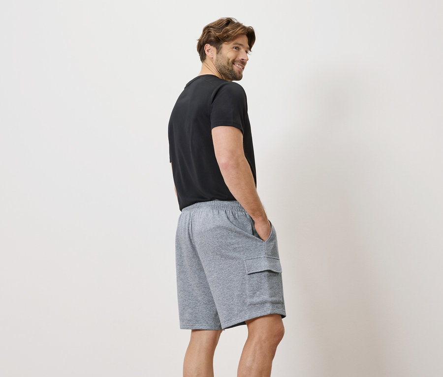 Mann trägt ein schwarzes T-Shirt und graue Shorts mit Cargotaschen, die Hände in den Hosentaschen, blickt über die Schulter.