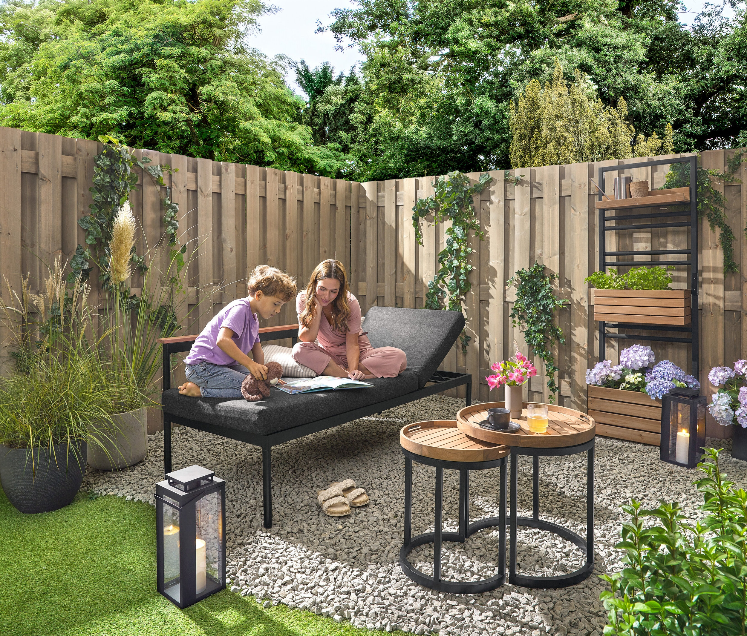 Ein Kind und eine Frau sitzen auf einer 2-in-1-Gartenbank ‒ Sonnenliege »Elin« und lesen ein Buch. Im Hintergrund befindet sich eine Pflanzwand mit flexibel einhängbaren Regalen »Elin«. Eine Outdoor-Laterne mit Kerze steht rechts. Zwei Garten-Beistelltische mit abnehmbaren Tabletts, Ø ca. 40 cm und 50 cm stehen auch da, sowie 2 Outdoor-Kissen mit Leinen-Haptik.