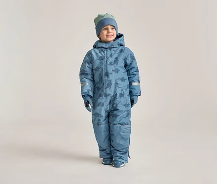 Ein lächelnder Junge trägt einen blauen Schneeanzug mit Dinosaurier-Muster, passende Handschuhe und eine Mütze vor einem hellen Hintergrund.