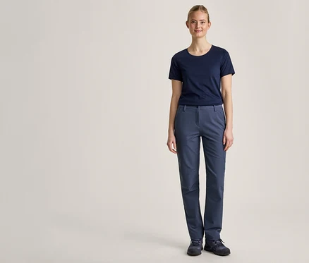 Eine junge Frau steht frontal vor einem hellen Hintergrund, trägt ein dunkelblaues T-Shirt, eine blaue Hose und dunkelblaue Schuhe.