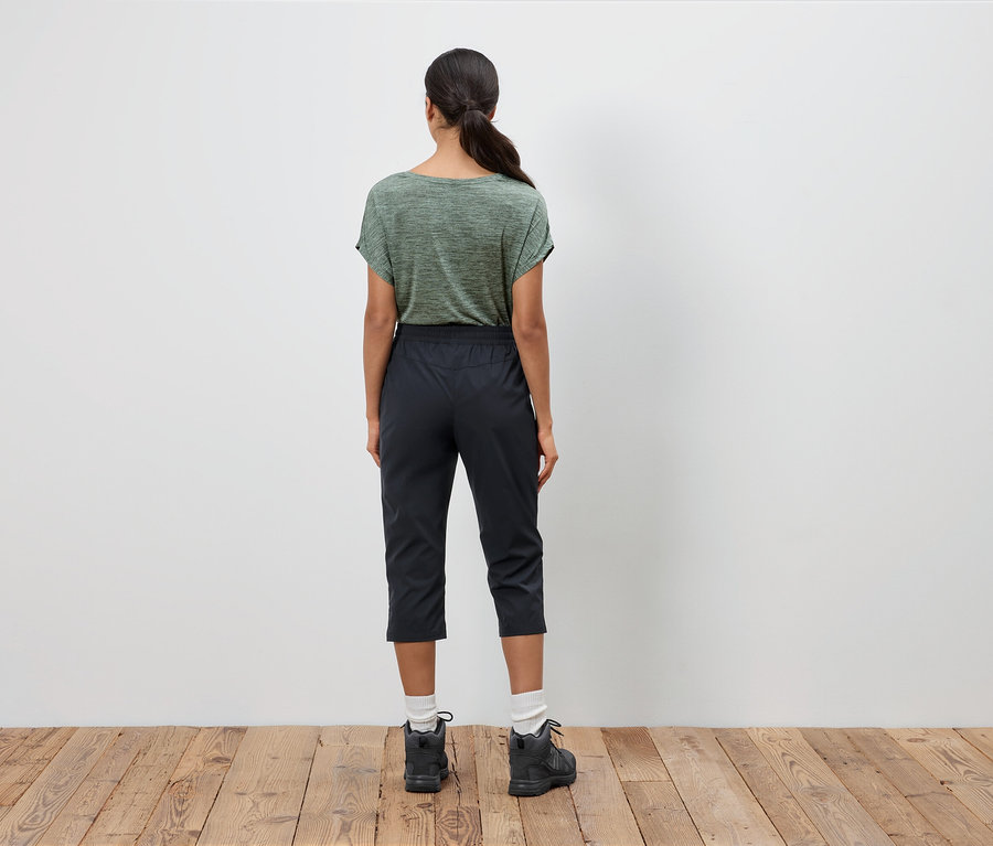 Rückansicht einer einzelnen Frau mit Pferdeschwanz, die ein olivgrünes T-Shirt, eine schwarze Dreiviertelhose und Wanderschuhe auf einem Holzboden trägt.