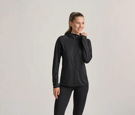Eine Frau in einer schwarzen Kapuzenjacke und schwarzen Leggings, lächelt und blickt nach rechts, ihre linke Hand an der Kapuze. Ihr Haar ist zu einem Pferdeschwanz gebunden.