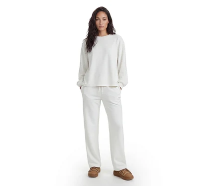 Frau steht in einem weißen Langarmshirt, passender weißer Jogginghose und braunen Hausschuhen.