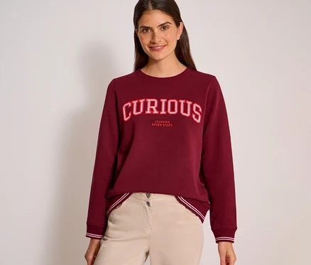 Eine lächelnde Frau mit dunklen Haaren trägt ein bordeauxrotes CECIL Sweatshirt mit der Aufschrift „CURIOUS“ in rot-weißen Buchstaben und „LEARNING NEVER STOPS“ darunter, kombiniert mit hellbeiger Hose.
