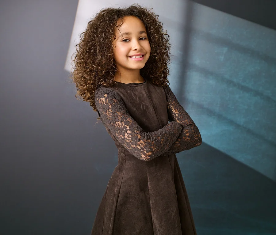 Lächelndes junges Mädchen mit lockigem braunem Haar trägt ein braunes Kleid mit Spitzärmeln und verschränkten Armen vor dunklem Hintergrund.